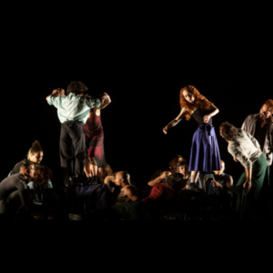 Companhia de Dança Palácio das Artes. Sobre o palco em fundo escuro, grupos de cerca de 10 pessoas realizam uma performance, com alguns em pé e outros agachados