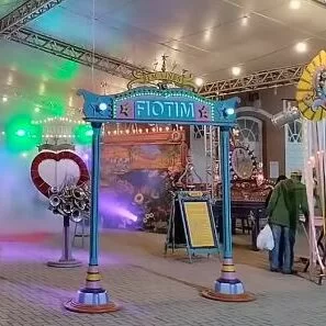 Pórtico azul do projeto Fiotim sobre um piso de tijolinhos cinza claro. Ao fundo, painéis impressos numa lona e um banner de chão azul com papel amarelo.