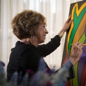 Eni D’Carvalho, de perfil, pinta uma tela. Eni é uma senhora de cabelo aloirados e curtos. Ela Tem brincos grande de argola e veste blusa preta.