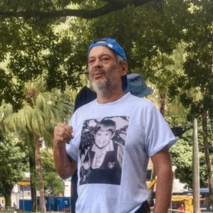 Dudu Melo é um homem branco, 60 mais, com barba grisalha. Veste boné azul virado pra trás e camiseta branca. Ao fundo, árvores e vegetação em um parque