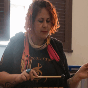 Josiana Dutra é uma mulher branca de cabelos vermelhos e curtos pouco acima dos ombros. Usa cachecol e segura uma baqueta.