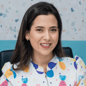 Virginia Machado é uma mulher branca, de cabelos pretos lisos na altura dos ombros. Veste blusa branca com estampas de abacaxis amarelos, rosas e azuis.